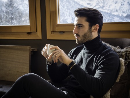 Young handsome man with beard drinking coffee or tea or hot chocolate, looking outside the window, away confidently on background of snowy mountains.の写真素材
