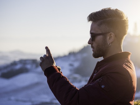 Handsome young man outdoor in winter fashion using smartphone to take photo of the landscape, wearing black coat and woolen scarfの写真素材