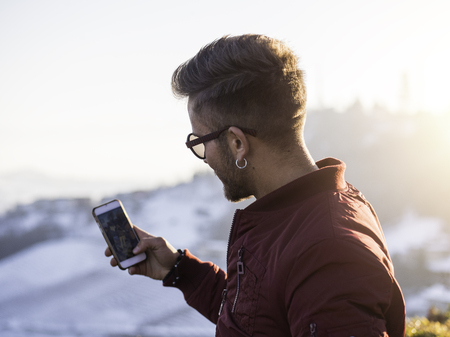 Handsome young man outdoor in winter fashion using smartphone to take photo of the landscape,...