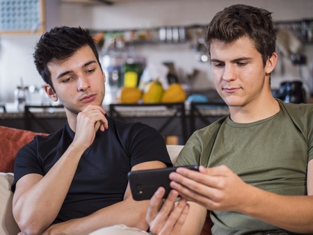 Two casual young guys relaxing on sofa at home and watching smartphone together.