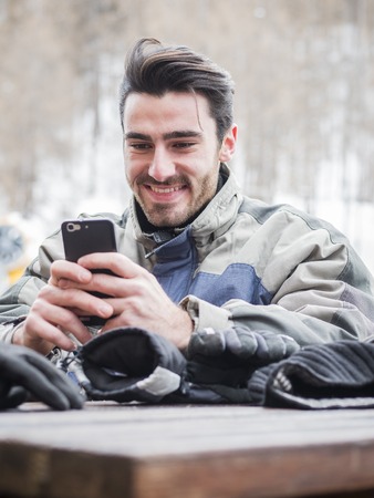 Handsome young man in skiing suit relaxing, typing text message or surfing the internet, sitting at...