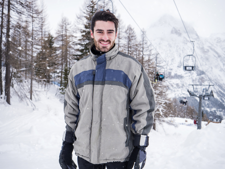 Young handsome man in winter sportswear looking at camera, up snow covered mountainsの写真素材