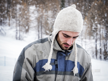 Young handsome man in winter sportswear and wool cap, looking away up a snowy mountain