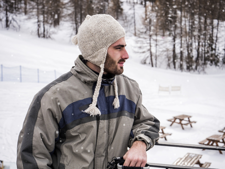 Young handsome man in winter sportswear and wool cap, looking away up a snowy mountainの写真素材