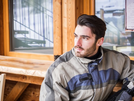 Handsome young man in skiing suit relaxing, sitting at table outside ski resortの写真素材