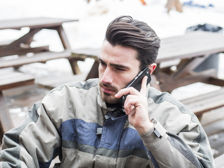 Handsome young man in skiing suit relaxing, using cell phone to make a call, sitting at table...