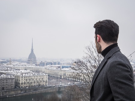 Handsome young man standing outside in winter, on balcony overlooking snowy cityscape or Turim, in Italyの写真素材