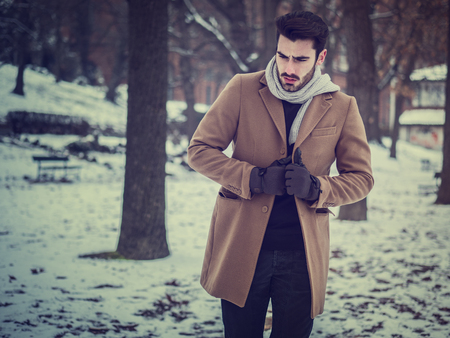 Handsome young man standing outside in winter, in snowy city park. Cold urban settingの写真素材