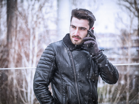 Handsome trendy man wearing black leather jacket standing and talking on cell phone, outdoor in city setting in winter day shotの写真素材