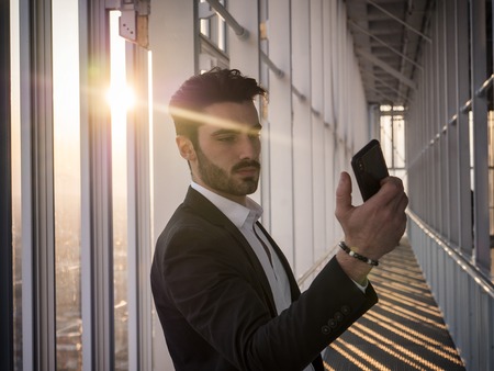 Handsome serious businessman standing inside modern building using cellphone next to big window, taking photo with camera phoneの写真素材