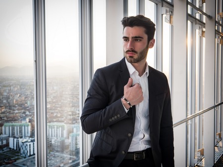 Handsome serious businessman standing inside modern building next to big window, looking out