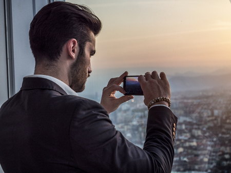 Handsome serious businessman standing inside modern building using cellphone next to big window, taking photo with camera phoneの写真素材