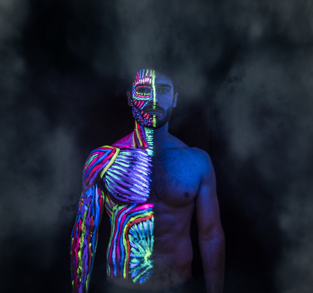 Young man painted in fluorescent paint on face and muscular torso, in studio shot with UV lightの写真素材