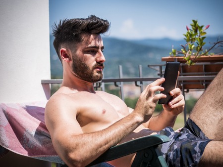 Shirtless Young Man Drying Off in Hot Sun Reading an Ebook with Electronic Reader, Muscular Man...