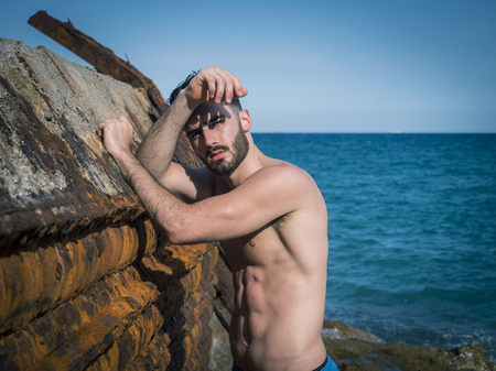 Handsome young man standing next to rusty metal structure, shirtless wearing boxer shorts. Looking at camera, with sea or ocean behindの写真素材