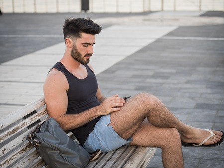 Young handsome man using smartphone, typing text message to someone while sitting on a bench at the...