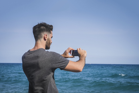 Full body shot of a handsome young man using cell phone to take photo, crouching on a beach, wearing boxer shorts and dark t-shirtの写真素材