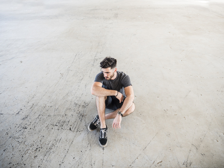 Fashionable Handsome Young Man Posing, Sitting on the Ground Inside an Empty Building While Looking...