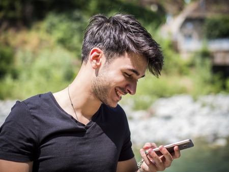 Young handsome man standing in park in autumn, using a cell phone to send text message, typing on it
