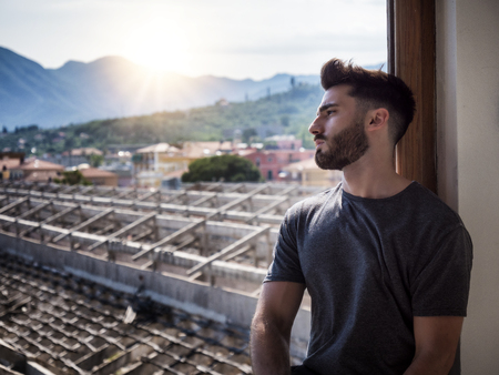 Handsome young man sitting by window, thoughtful and sad, in a summer day