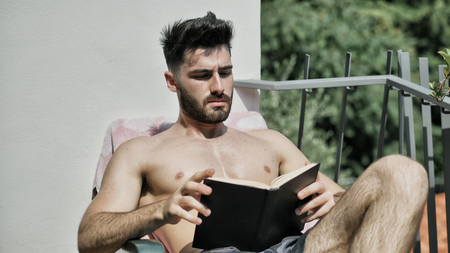 Shirtless Young Man Drying Off in Hot Sun Reading a Book, Muscular Man Wearing Bathing Suit Sunbathing on Beach Lounge Chairの写真素材