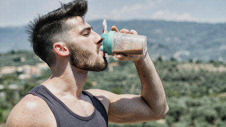 Young muscular shirtless man drinking protein shaker from blender on a balcony in summerの写真素材