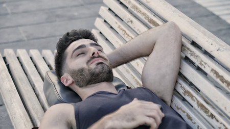 Attractive young man resting on wood bench outdoor in city park during the eveningの写真素材
