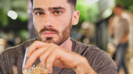 Young handsome man drinking cocktail outside at night sitting at bar table in a summer day in city