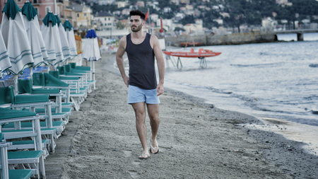 Attractive fit athletic young man walking along the beach among lounge chairs and sun umbrellas, wearing black tank-topの写真素材