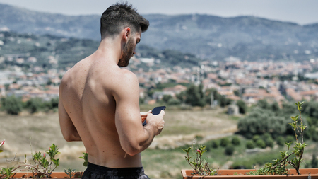 Young handsome man using smartphone, typing text message to someone while standing shirtless on a balcony at the seaside over countryside landscapeの写真素材