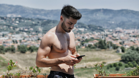 Young handsome man using smartphone, typing text message to someone while standing shirtless on a balcony at the seaside over countryside landscapeの写真素材