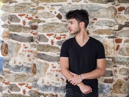 Handsome young man standing in European city street, leaning against a wall looking away to a side