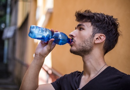 Handsome young man drinking water from plastic bottle, standing in European city street, leaning against a wallの写真素材