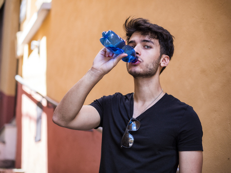 Handsome young man drinking water from plastic bottle, standing in European city street, leaning against a wall looking at cameraの写真素材