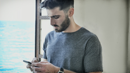 Athletic young man at the seaside using cell phone to type message while looking at the sea and beachの写真素材