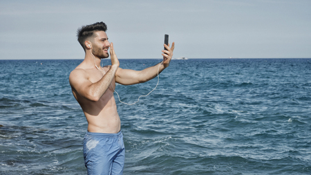 Young shirtless muscular handsome man doing videochat with smartphone by the sea with earphonesの写真素材