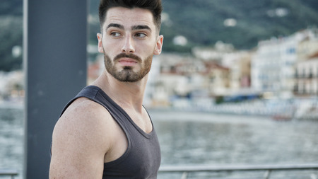 Attractive, sad, troubled, worried young man at sunset on seaside boardwalk or seafront, wearing black tank-top, looking at the sea or oceanの写真素材