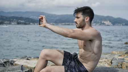 Young handsome man at the seaside using smartphone to take shots and film footage of the sea or oceanの写真素材