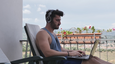 Waist Up Profile of Attractive Man with Dark Hair, Sitting with Laptop Computer Listening to Music with Big Headphonesの写真素材