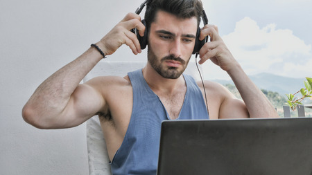 Waist Up Profile of Attractive Man with Dark Hair, Sitting with Laptop Computer Listening to Music with Big Headphonesの写真素材