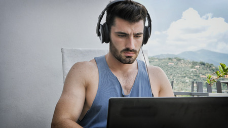 Waist Up Profile of Attractive Man with Dark Hair, Sitting with Laptop Computer Listening to Music with Big Headphonesの写真素材