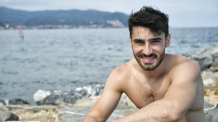 Attractive fit athletic young man soaking in the sun on seaside boardwalk or seafront, sitting on rock, shirtless, looking at camera with a smileの写真素材