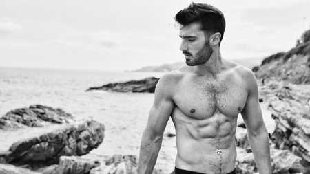 Attractive fit athletic young man soaking in the sun at rocky beach in front of the sea, shirtlessの写真素材