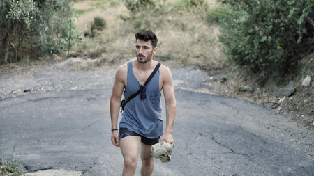 Handsome young man outdoor hiking on rural road with backpack on shoulder, looking at country around himの写真素材