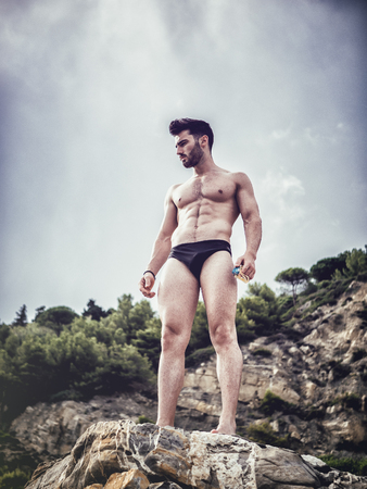 Handsome muscular young man standing against blue sky, relaxed, shirtless, in a majestic pose seen from belowの写真素材