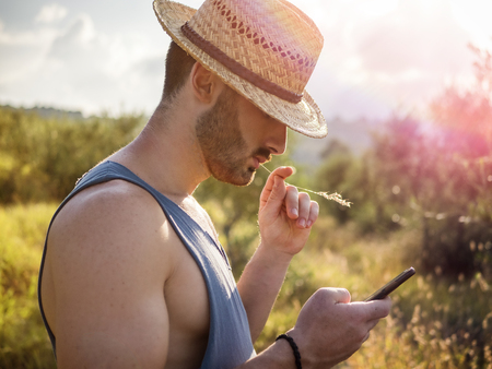 Attractive, fit handsome male farmer outdoor with cell phone, wearing straw hatの写真素材