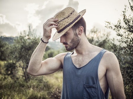 Attractive, fit young farmer man relaxing walking in a grass field, wearing straw hatの写真素材