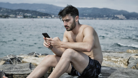 Solitary fit young man sitting on a stone at the beach while listening to music through earphonesの写真素材