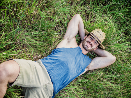 Attractive, fit young man relaxing laying down in a grass field, wearing straw hat, looking at camera, smilingの写真素材
