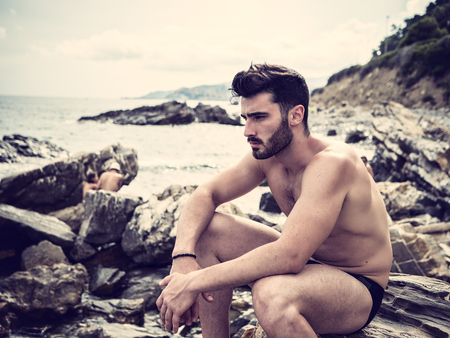 Handsome muscular young man sitting on a rocky beach, relaxed, shirtless, looking at cameraの写真素材